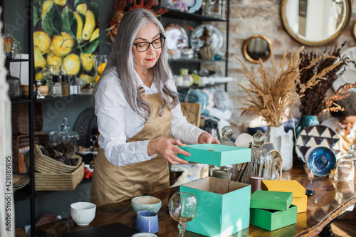 Attractive aged woman in eyeglasses and apron doing inventory at decor store, opening green box. Competent saleswoman rechecking all available assortment at shop. photo
