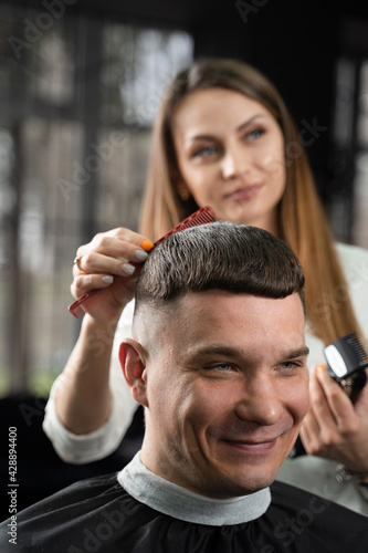 Woman barber making hairstyle in barbershop using clipper. Hairdresser cutting hair of handsome man.