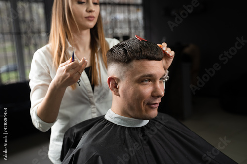 Woman barber making hairstyle in barbershop using clipper. Hairdresser cutting hair of handsome man.