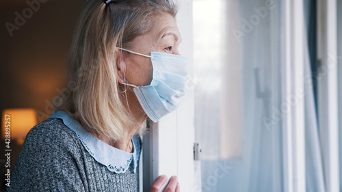 Portrait of elderly woman with medical face mask in quarantine looking through the window. High quality photo