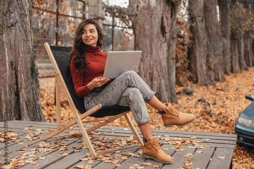 Beautiful woman in cozy outfit works at laptop while sitting on chair in autumn park.