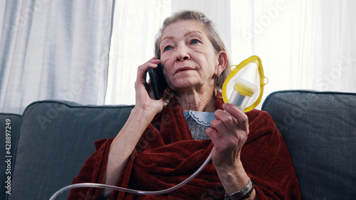 Vulnerable old woman having a phone call and using inhaler. Difficulty breathing at old age. High quality photo