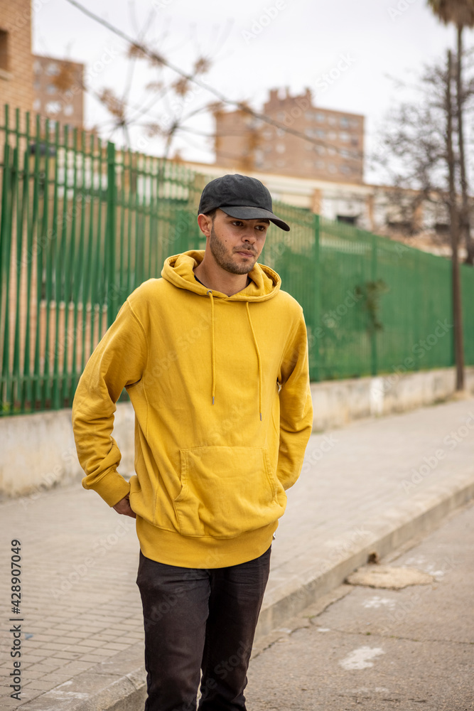 Young caucasian male in a yellow hoodie and black cap standing on the street Young caucasian male in a yellow hoodie and black cap standing on the street