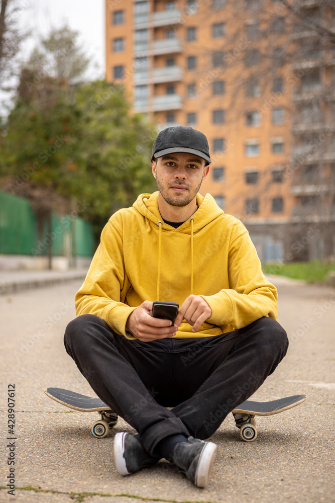 Obraz premium Young caucasian male from Italy in a yellow hoodie and black cap sitting on a skateboard with a smartphone