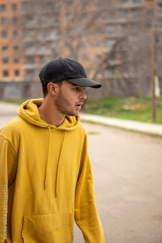 Young caucasian male in a yellow hoodie and black cap standing on the street Young caucasian male in a yellow hoodie and black cap standing on the street