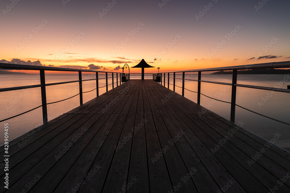 Incredible long exposure of a pier with a vanishing point perspective. Wooden board that leads to a parasol and a bell at its end. Sea and sky in pastel colors of orange, yellow and pink.