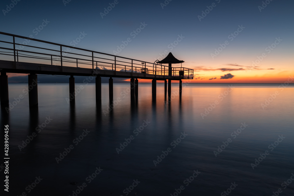 Incredible long exposure of a pier with a vanishing point perspective. Wharf with some colums over the smooth sea. Pastel tones of orange, yellow, blue and pink.