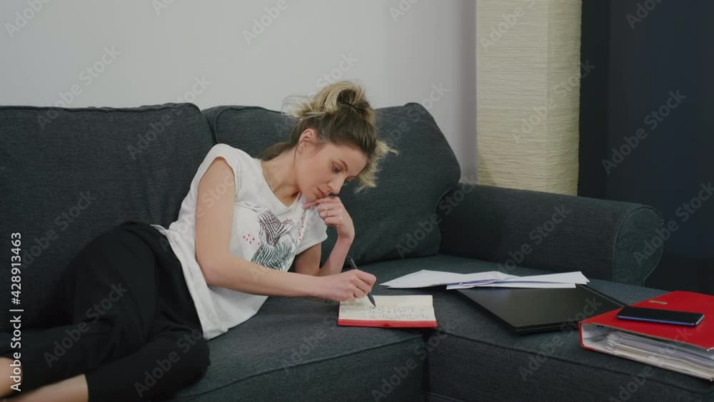 Pretty woman makes notes in notebook while lying on couch at home in a modern apartment. Is preparing for important meeting. Very focused prepares the speech. Write with a pen in your notebook. Pretty woman makes notes in notebook while lying on couch at home in a modern apartment. Is preparing for important meeting. Very focused prepares the speech. Write with a pen in your notebook.