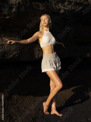 Attractive Caucasian woman posing on the beach. Woman standing near the rock. Sexy slim body. Summer concept. Exotic country travel concept. Lifestyle. Black sand beach in Bali