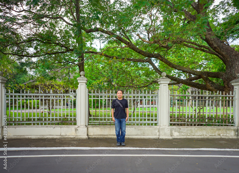Asian Black T-Shirt is standing in front of white vintage fence beside the road in the afternoon time. Asian Black T-Shirt is standing in front of white vintage fence beside the road in the afternoon time.