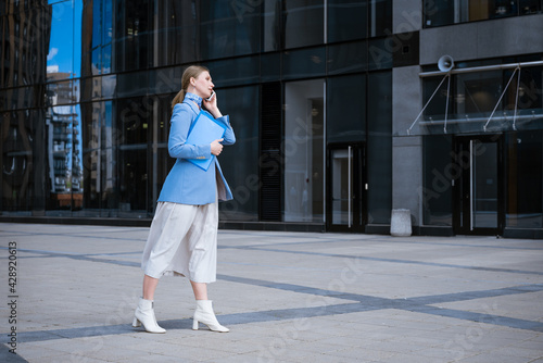 caucasian business woman in a blue jacket and dress talking on the phone with a folder of papers in her hand against the background of an office building