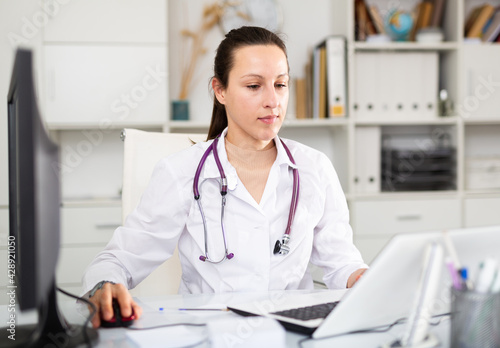 Young female therapist working in a clinic is sitting at her workplace in an office, typing on a computer