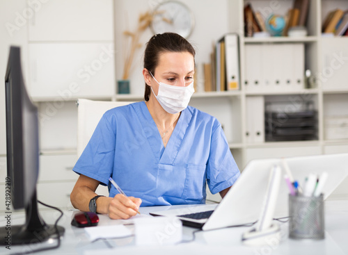 Young woman doctor in a protective mask working in a clinic writes important notes while sitting at a work desk in ..front of a computer