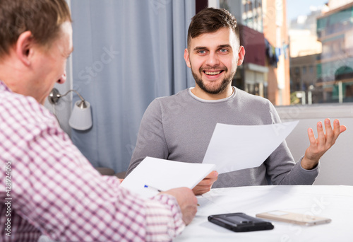 Happy man discussing papers with male colleague while sitting at desk in home interior