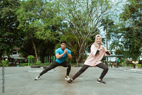 a young man and a veiled girl in gym clothes doing the leg warm-up movement together before exercising in the park