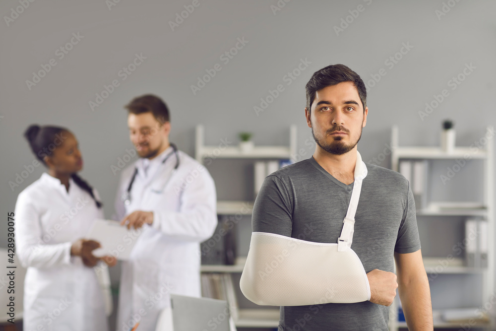 Medicine and healthcare. Serious man patient with broken arm wrapped on orthopedic bandage to immobilize limb after injury. Medic doctor team on blurred background. Headshot portrait with copy space