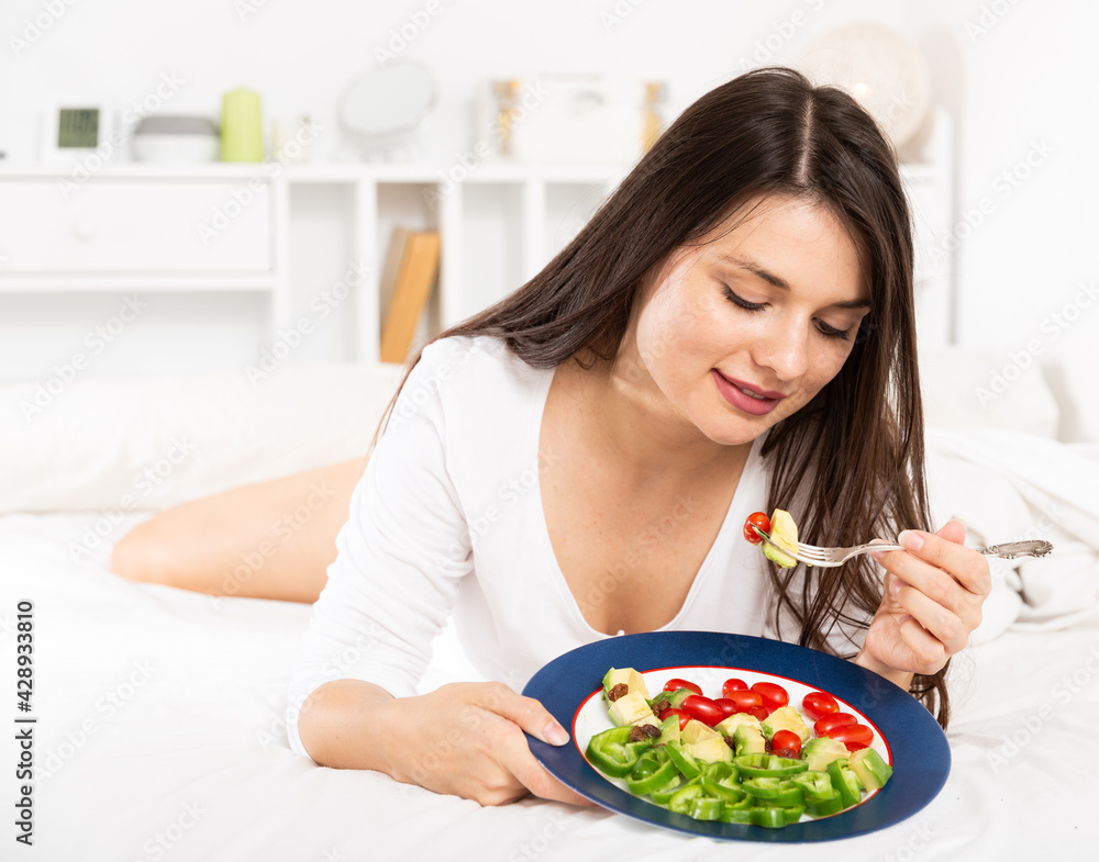 Pretty woman in white dress holding fork and eating vegetable salad in bedroom