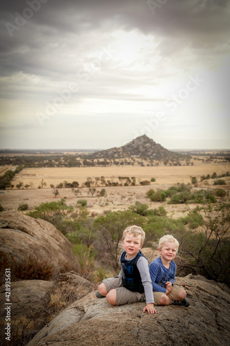 Vertical portrait image of little boys sitting on rock during bushwalk with view of Pyramid Hill, Victoria Australia in the background. Nature walk on a cloudy day.