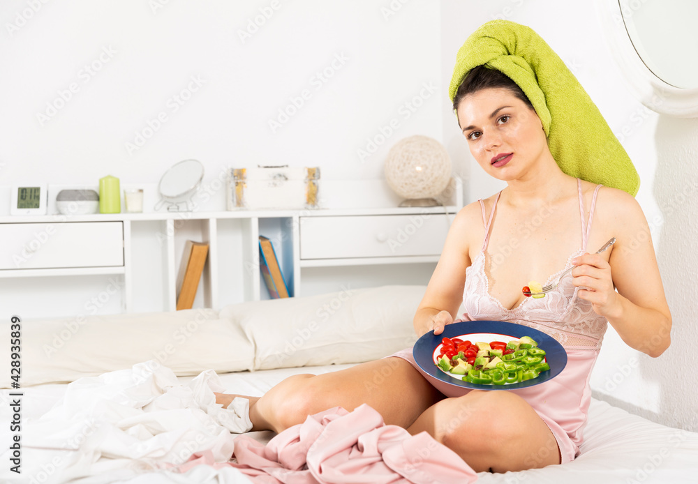 Young pretty woman with a towel on his head eating vegetable salad from plato in bed at home
