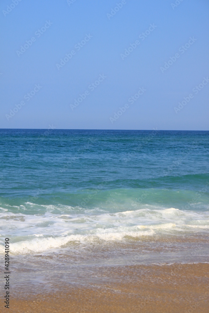La mer le ciel sont bleus, le sable fin est jaune, quelques vagues se dessinent sur l´eau devenant verte proche de la plage.