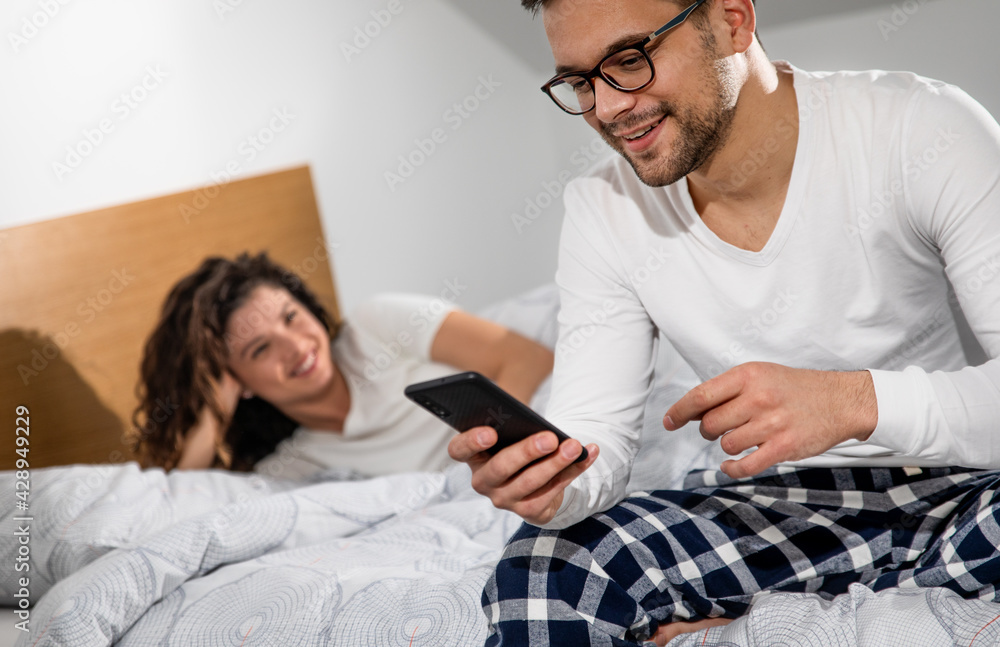 Obraz premium Man using phone in bed while woman looking at him.