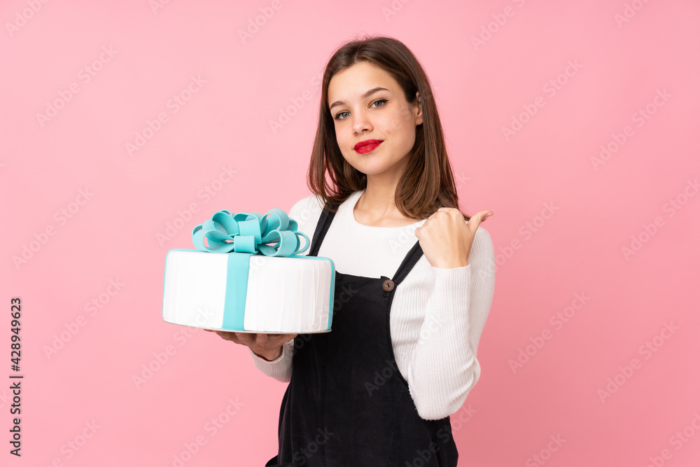 Girl holding a big cake isolated on pink background pointing to the side to present a product Girl holding a big cake isolated on pink background pointing to the side to present a product