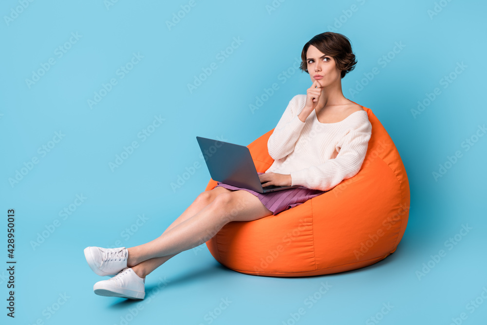 Full length photo portrait of thoughtful girl with laptop sitting in orange beanbag isolated on pastel blue colored background Full length photo portrait of thoughtful girl with laptop sitting in orange beanbag isolated on pastel blue colored background