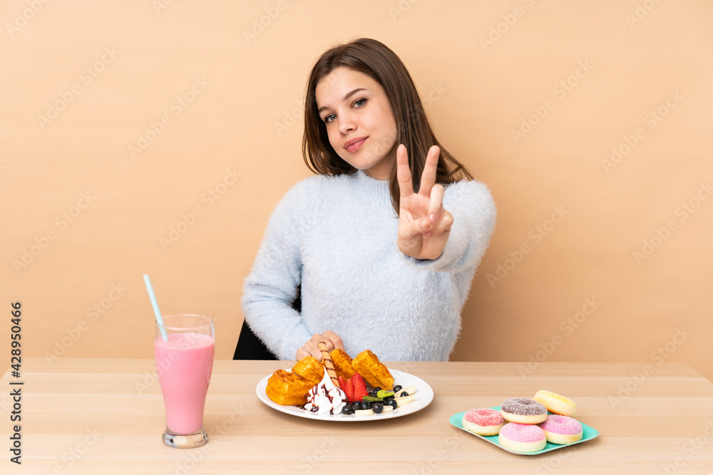 Teenager girl eating waffles isolated on beige background smiling and showing victory sign Teenager girl eating waffles isolated on beige background smiling and showing victory sign