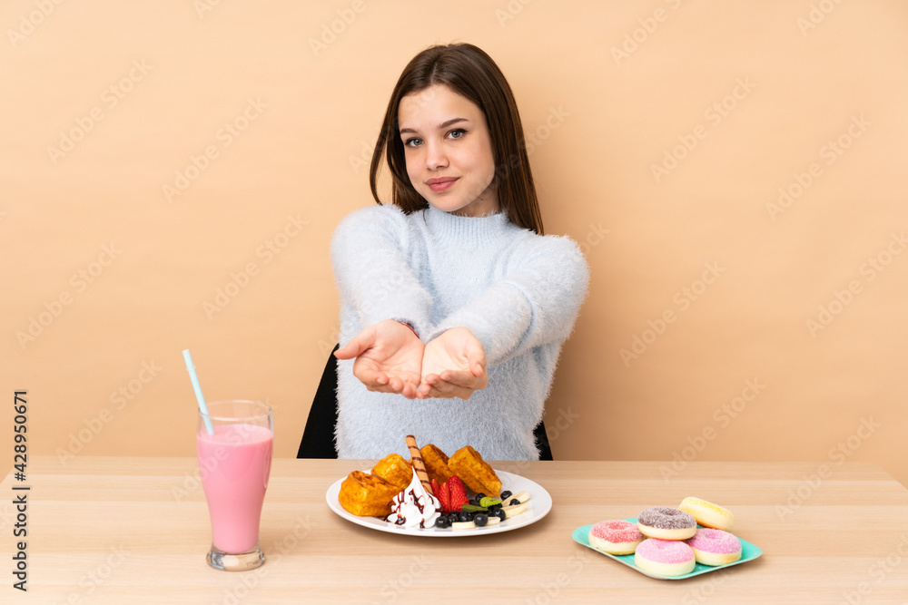 Teenager girl eating waffles isolated on beige background holding copyspace imaginary on the palm to insert an ad Teenager girl eating waffles isolated on beige background holding copyspace imaginary on the palm to insert an ad