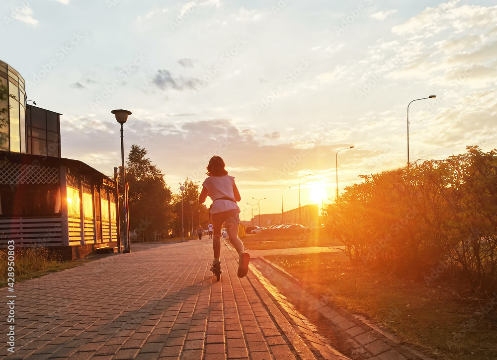 A teenage girl rides a scooter on the city sidewalk in the rays of the setting sun