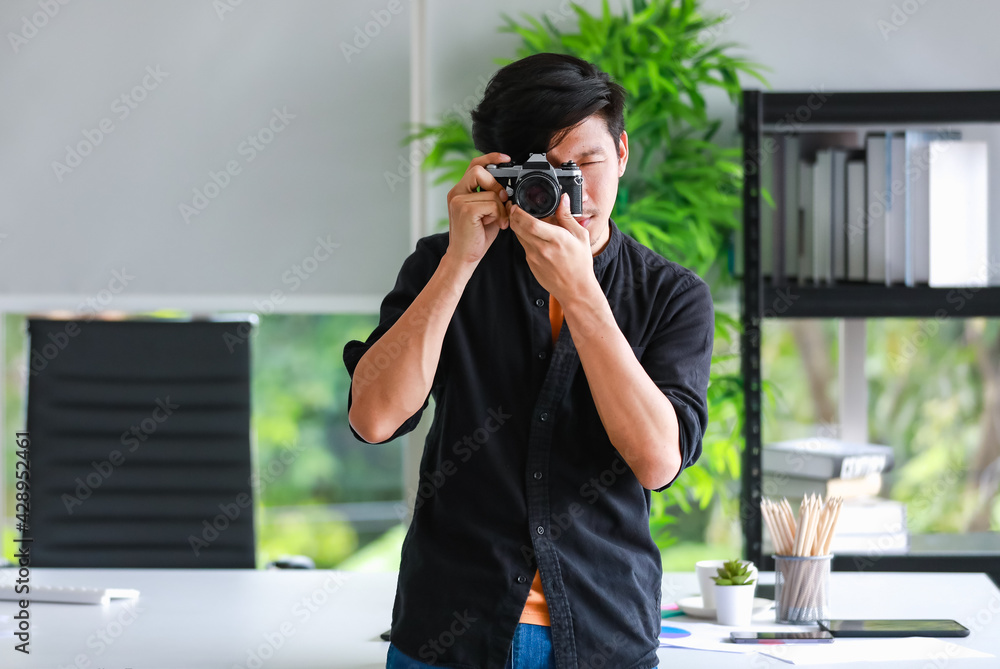 A trendy professional photographer standing against working desk in the office, holding and testing a retro camera, by taking pictures to checking the new features A trendy professional photographer standing against working desk in the office, holding and testing a retro camera, by taking pictures to checking the new features