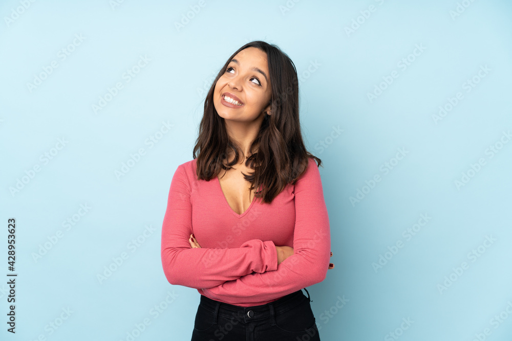 Young mixed race woman isolated on blue background looking up while smiling Young mixed race woman isolated on blue background looking up while smiling