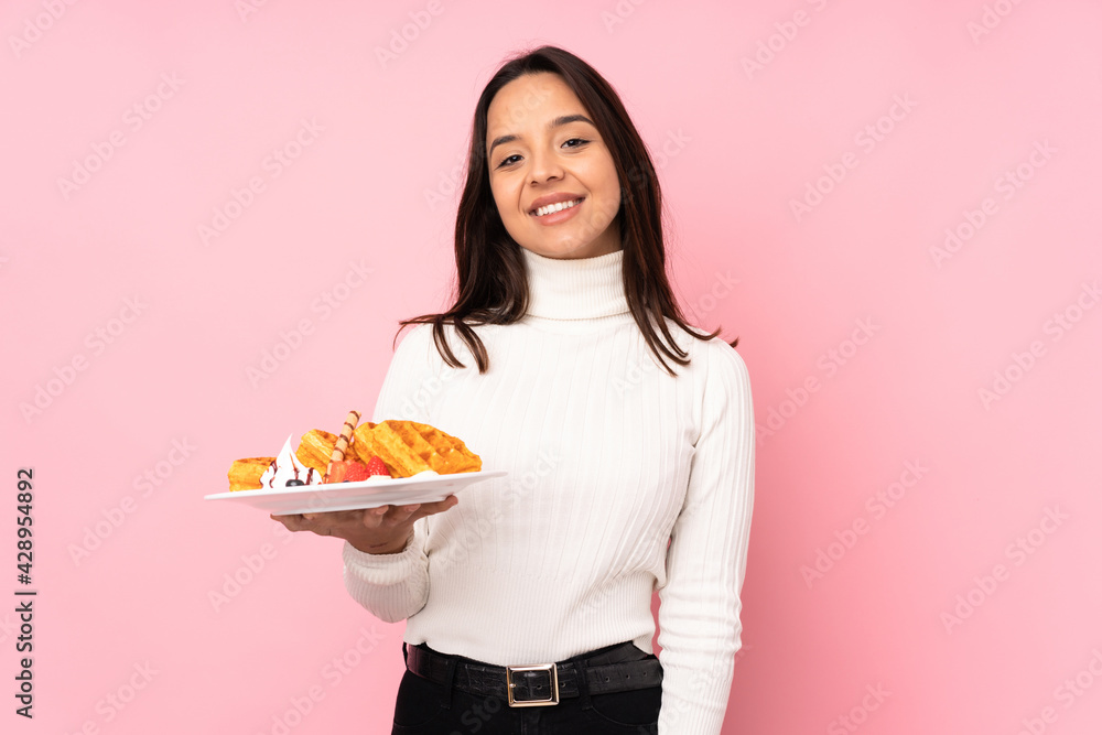Young brunette woman holding waffles over isolated pink background laughing