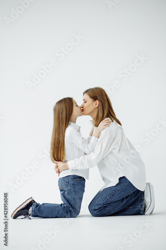 Mother's Day . Mom and daughter on their knees kissing each other on a white background, wearing white shirts and jeans, side view.