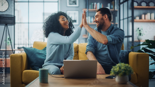 Diverse Multiethnic Couple are Sitting on a Couch Sofa in Stylish Living Room and Choosing Items to Buy Online with Laptop Computer, Give High Five. Friends or Colleagues are Discuss Work Projects. photo