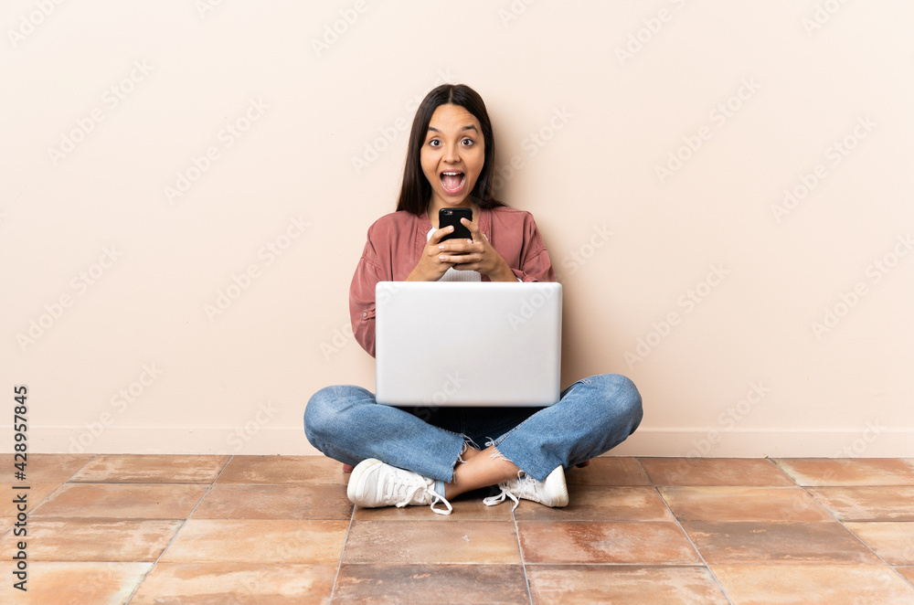 Young mixed race woman with a laptop sitting on the floor surprised and sending a message Young mixed race woman with a laptop sitting on the floor surprised and sending a message