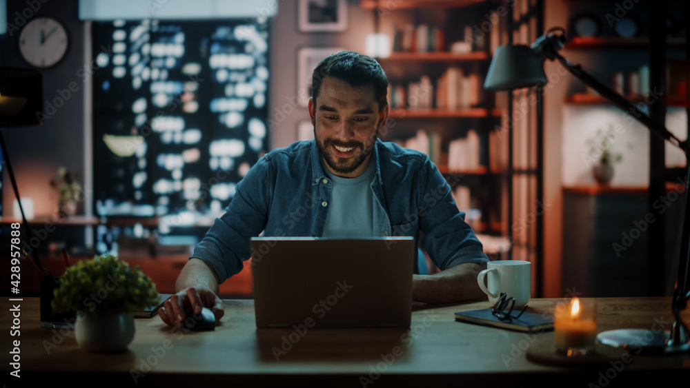 Handsome Caucasian Man Working on Laptop Computer while Sitting at a Desk in Dark Cozy Living Room in the Evening. Freelancer Working From Home. Browsing Internet, Using Social Networks, Having Fun. Handsome Caucasian Man Working on Laptop Computer while Sitting at a Desk in Dark Cozy Living Room in the Evening. Freelancer Working From Home. Browsing Internet, Using Social Networks, Having Fun.