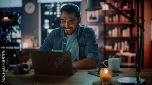 Handsome Caucasian Man Working on Laptop Computer while Sitting at a Desk in Dark Cozy Living Room in the Evening. Freelancer Working From Home. Browsing Internet, Using Social Networks, Having Fun.