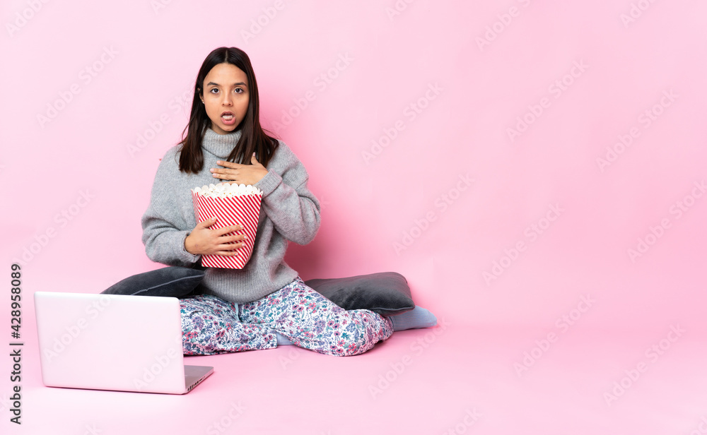 Young mixed race woman eating popcorn while watching a movie on the laptop surprised and shocked while looking right