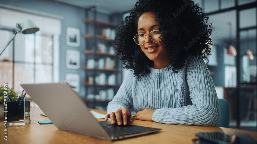 Beautiful Authentic Latina Female with Afro Hair Sitting at a Desk in a Cozy Living Room and Using Laptop Computer at Home. She's Browsing the Internet and Checking Videos on Social Networks. Beautiful Authentic Latina Female with Afro Hair Sitting at a Desk in a Cozy Living Room and Using Laptop Computer at Home. She's Browsing the Internet and Checking Videos on Social Networks.