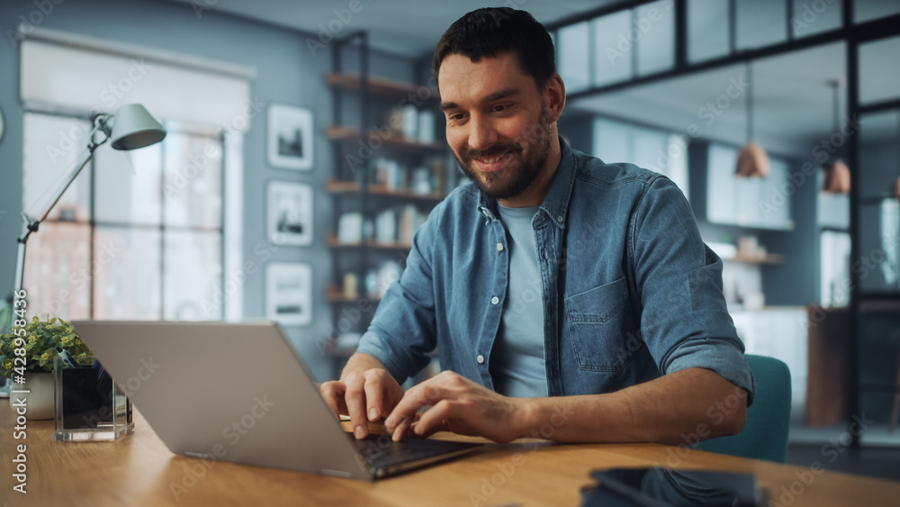 Handsome Caucasian Man Working on Laptop Computer while Sitting on a Sofa Couch in Stylish Cozy Living Room. Freelancer Working From Home. Browsing Internet, Using Social Networks, Having Fun. Handsome Caucasian Man Working on Laptop Computer while Sitting on a Sofa Couch in Stylish Cozy Living Room. Freelancer Working From Home. Browsing Internet, Using Social Networks, Having Fun.