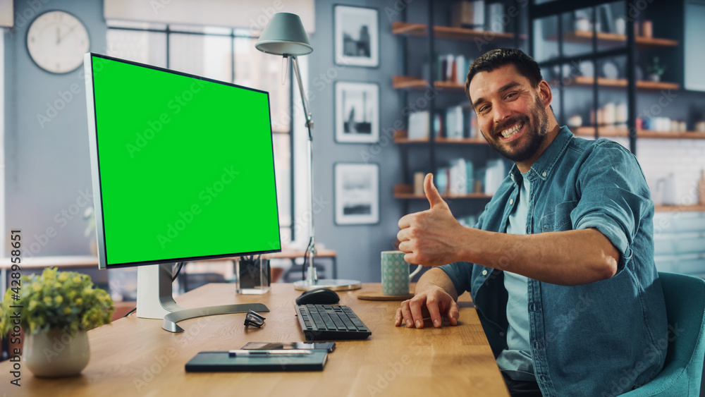 Handsome Caucasian Specialist Working on Desktop Computer with Green Screen Mock Up Display at Home Living Room. Happy Freelance Man Smiles to the Camera and Shows a Thumbs Up Hand Gesture. Handsome Caucasian Specialist Working on Desktop Computer with Green Screen Mock Up Display at Home Living Room. Happy Freelance Man Smiles to the Camera and Shows a Thumbs Up Hand Gesture.