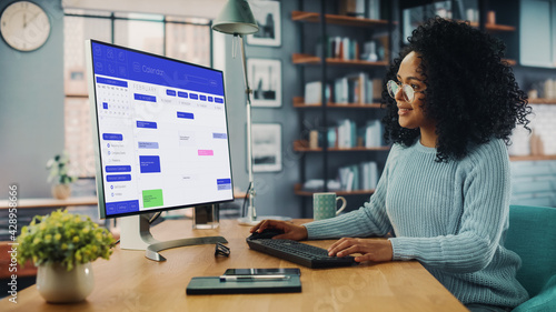 Latina Female Specialist Working on Desktop Computer at Home Living Room while Sitting at a Table. Freelancer Female Checking Her Calendar to Make an Appointment with New Clients and Employer.