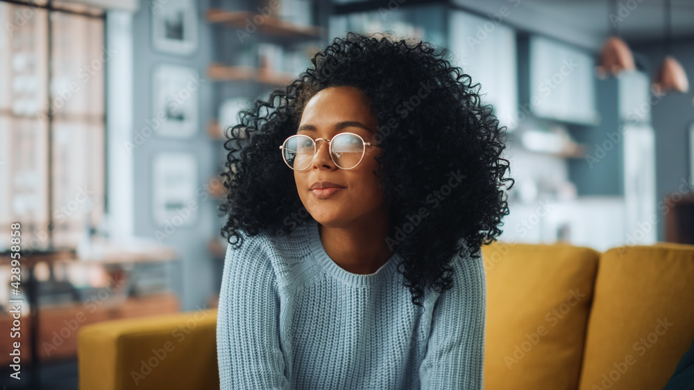 © Gorodenkoff - Portrait of a Beautiful Authentic Latina Female with Afro Hair Wearing Light Blue Jumper and Glasses. She Looks Away and Thinking about Life. Successful Woman Resting in Bright Living Room.