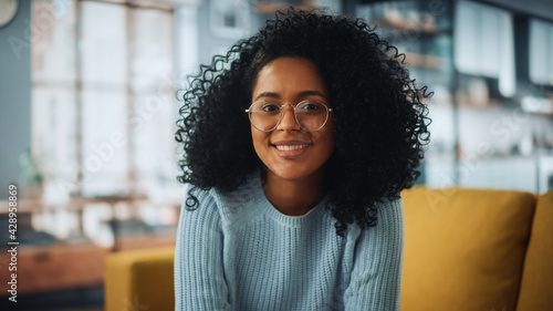 Portrait of a Beautiful Authentic Latina Female with Afro Hair Wearing Light Blue Jumper and Glasses. She Looks to the Camera and Smiling Charmingly. Successful Woman Resting in Bright Living Room. photo