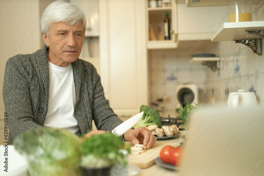 Serious concentrated retired man cooking dinner in cozy kitchen slicing fresh ingredients on cooking board, watching football game or series online on open laptop, having focused facial expression Serious concentrated retired man cooking dinner in cozy kitchen slicing fresh ingredients on cooking board, watching football game or series online on open laptop, having focused facial expression