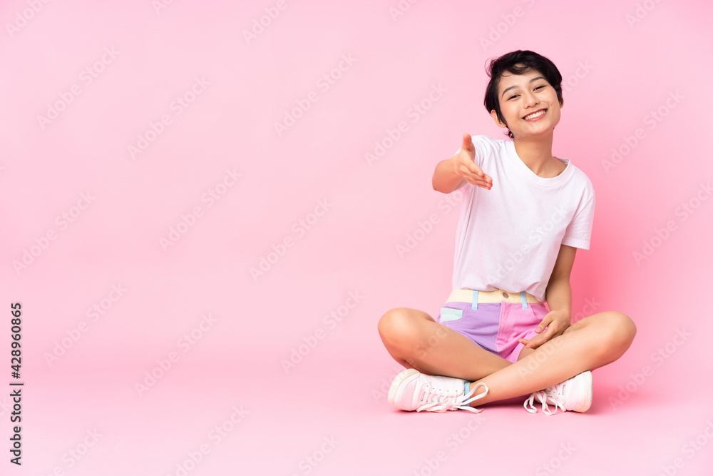 Young Vietnamese woman with short hair sitting on the floor over isolated pink background shaking hands for closing a good deal Young Vietnamese woman with short hair sitting on the floor over isolated pink background shaking hands for closing a good deal