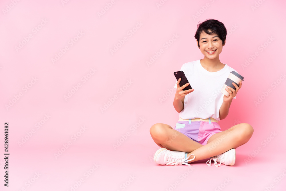 Young Vietnamese woman with short hair sitting on the floor over isolated pink background holding coffee to take away and a mobile Young Vietnamese woman with short hair sitting on the floor over isolated pink background holding coffee to take away and a mobile
