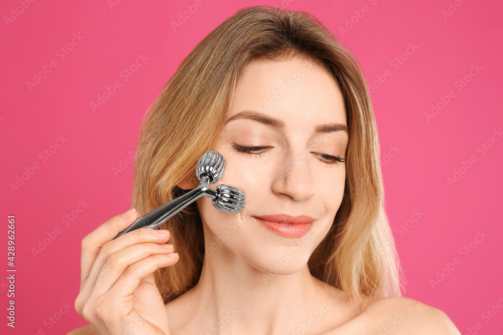 Young woman using metal face roller on pink background Young woman using metal face roller on pink background