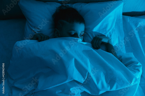 Small boy lies in bed in dark night, afraid of nightmares and terrible dreams in children. Concept of fear and domestic violence